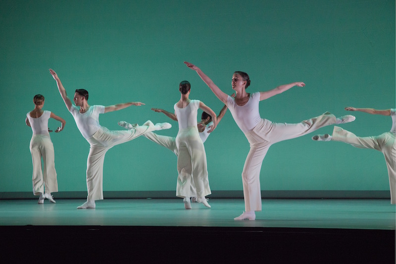 Dancers all in white execute expansive arabesques in front of a sea green background